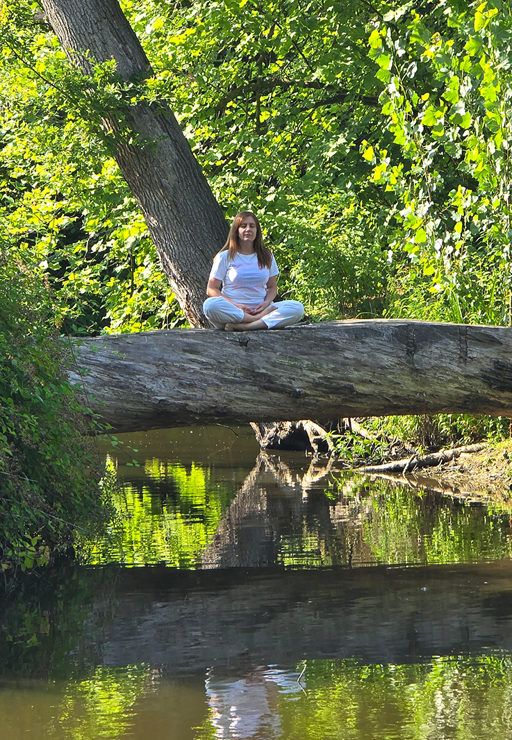 A woman meditating cross-legged on a fallen tree trunk over a stream surrounded by green foliage.